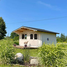 Load image into Gallery viewer, Front view of a fully assembled 12x15 Clifton Cabin with a covered terrace and double doors, surrounded by landscaped greenery and a clear blue sky by WholeWoodcabins