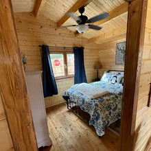 Load image into Gallery viewer, Cozy bedroom inside the cabin with wooden walls and ceiling, featuring a bed, windows, and natural light creating a warm, rustic atmosphere by WholeWoodCabins