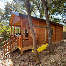 Load image into Gallery viewer, Side view of the Mountaineer 500 cabin showcasing the wooden porch and stairs, with a yellow swing hanging between two trees, set in a wooded area by WholeWoodCabins