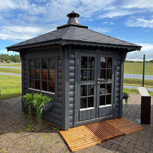 Load image into Gallery viewer, A dark gray wooden BBQ Pavilion with a pitched roof, double glass doors, and large windows, shown outdoors on a paved area with a wooden ramp in front. A grassy field and fence are visible in the background under a blue sky.
