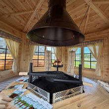 Load image into Gallery viewer, A black metal outdoor fireplace insert with a hanging conical hood and a small copper chimney, displayed on a wooden table inside a rustic log cabin with windows and natural light.