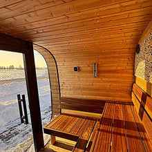 Load image into Gallery viewer, A wooden sauna interior with tiered wooden benches, a wood-paneled wall, a glass door opening onto snow outdoors, and a decorative wood-patterned wall section on the right.