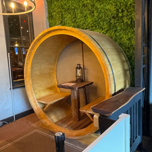Load image into Gallery viewer, Wooden barrel-shaped booth with seating and a small table inside, against a green wall.