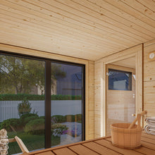 Load image into Gallery viewer, Interior view of a wooden sauna with a bench, bucket, and large glass doors overlooking a landscaped garden and modern house.