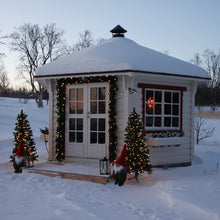 Load image into Gallery viewer, A snow-covered white BBQ Pavilion decorated for Christmas, with a snow-laden roof, white double doors framed by green garlands and warm string lights, a window with red trim, two small decorated Christmas trees, and gnomes in a snowy outdoor setting.