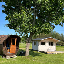 Load image into Gallery viewer, 12x15 Clifton Cabin with light natural wood finish and front terrace, placed in a sunny green yard next to a barrel sauna under a large tree by WholeWoodCabins
