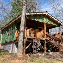 Load image into Gallery viewer, Wooden cabin with green siding and natural wood accents, featuring a front porch with stairs leading to the entrance, surrounded by trees by WholeWoodCabins
