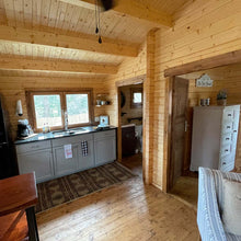 Load image into Gallery viewer, Cabin interior showing the kitchen area with wooden walls, cabinets, and large windows providing natural light, creating a functional yet rustic feel by WholeWoodCabins
