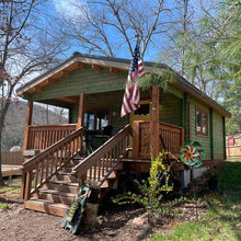 Load image into Gallery viewer, Front view of the cabin with green siding, wooden porch, and an American flag displayed at the entrance, surrounded by trees and garden decor by WholeWoodCabins
