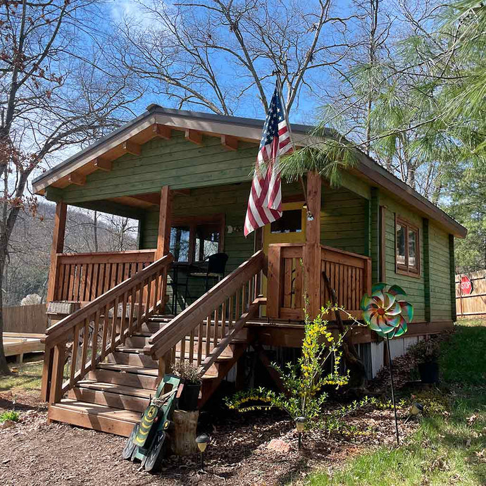 Front view of the cabin with green siding, wooden porch, and an American flag displayed at the entrance, surrounded by trees and garden decor by WholeWoodCabins