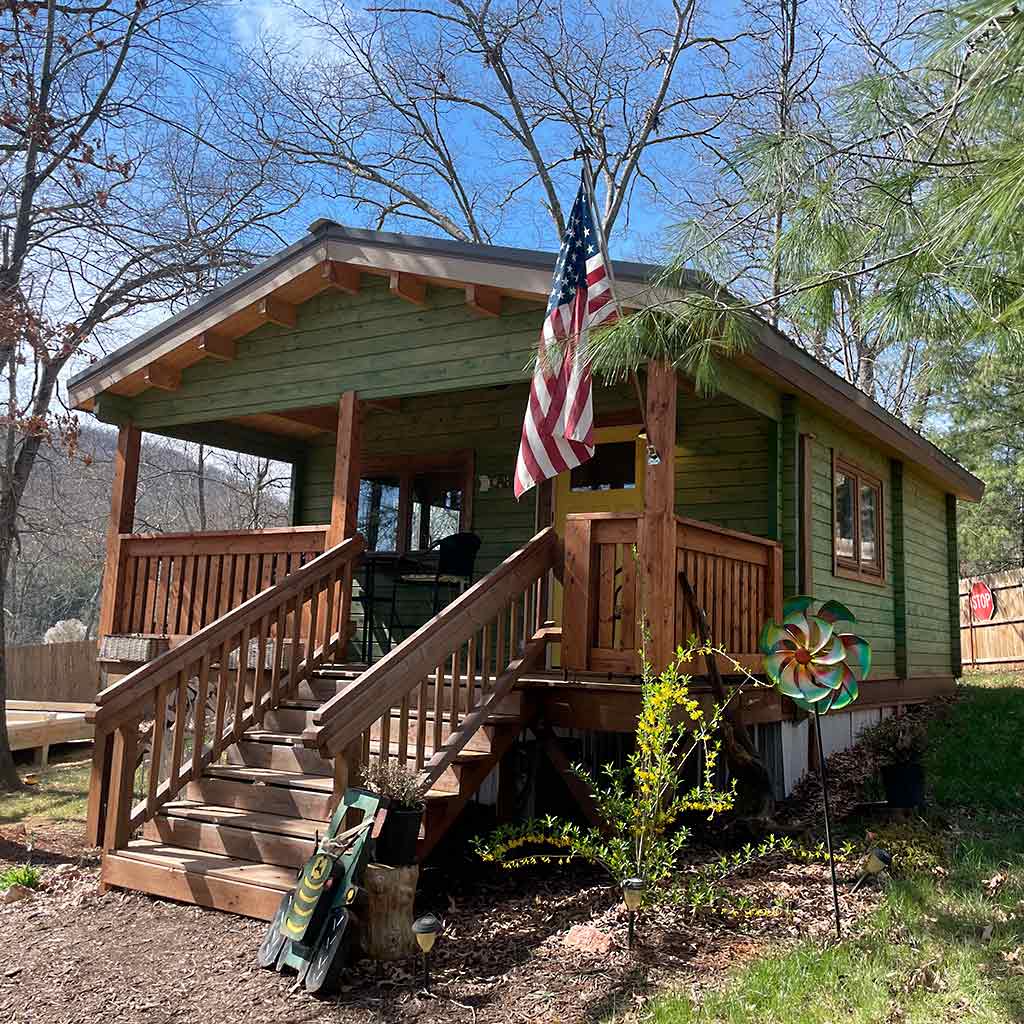 Front view of the cabin with green siding, wooden porch, and an American flag displayed at the entrance, surrounded by trees and garden decor by WholeWoodCabins