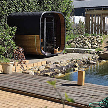 Load image into Gallery viewer, A modern outdoor cube shape sauna with a black roof and wood-paneled side, situated beside a stone-and-water pond with a wooden deck in the foreground.
