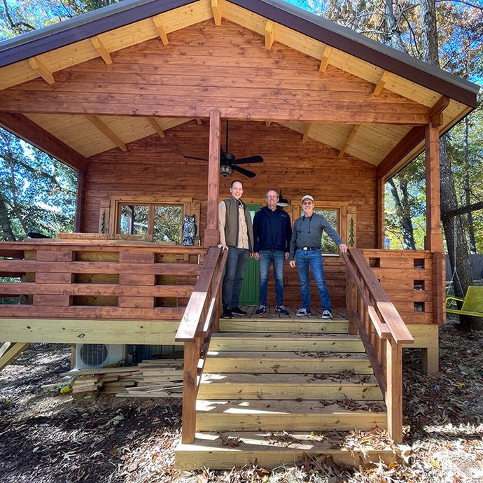 Front view of the Mountaineer 500 cabin with three people standing on the porch - WholeWoodCabins eam. The cabin features natural wood siding, exposed beams, and a wooden staircase by WholeWoodCabins