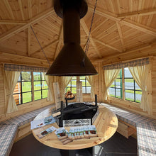 Load image into Gallery viewer, A rustic wooden chalet-style outdoor dining pavilion with a round wooden table, a black metal grill, built-in bench seating with plaid cushions, and a large central chimney stack under a pitched wooden roof.
