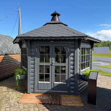 Load image into Gallery viewer, A small log BBQ pavilion with gray painted log walls, double glass doors, side windows, a dark gray shingled roof, and a copper-toned finial, shown on a paved area under a blue sky.
