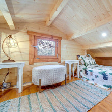 Load image into Gallery viewer, Wooden cabin loft interior with a bed, ottoman, and small table.
