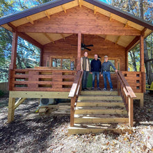 Load image into Gallery viewer, Front view of the Mountaineer 500 cabin with three people standing on the porch - WholeWoodCabins team. The cabin features natural wood siding, exposed beams, and a wooden staircase by WholeWoodCabins
