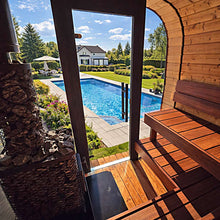 Load image into Gallery viewer, A wooden sauna interior with tiered seating and a stone-burned stove, overlooking a backyard pool, lawn, trees, and a house through an open doorway.
