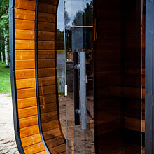 Load image into Gallery viewer, A modern wooden sauna with a curved exterior featuring warm brown wood panels and a large dark tinted glass door with a long metal handle. The sauna is situated outdoors near a sandy path, grass, and trees.
